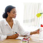 A woman sitting at a table with a notebook, planning and organizing with healthy snacks and water nearby. This image illustrates prep tips to keep in mind before breast pumping, emphasizing the importance of hydration, nutrition, and planning for a successful pumping session.