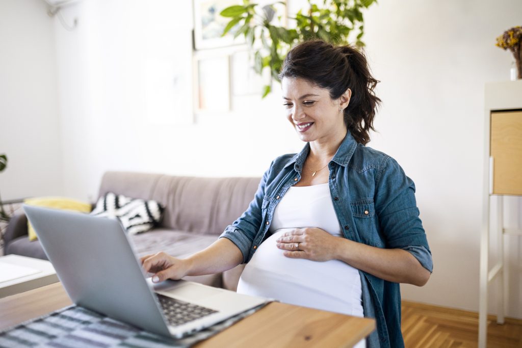 A pregnant woman smiling while researching on her laptop, possibly reading about breast pumps. This image represents breaking down Willow Pumps and exploring features to help expectant mothers make informed decisions.