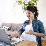 A pregnant woman smiling while researching on her laptop, possibly reading about breast pumps. This image represents breaking down Willow Pumps and exploring features to help expectant mothers make informed decisions.