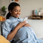 A pregnant woman smiling while looking at her phone, possibly researching Medela Breast Pumps. This image reflects the excitement and preparation for motherhood, including choosing the right breast pump.