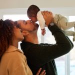 Mother with dark curly hair looking at her husband as he lifts and kisses their infant