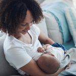 African American woman breastfeeding her infant on a couch after giving birth while contemplating how soon to pump or breastfeed