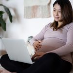 Pregnant woman in purple shirt sitting on bed looking at laptop ordering a breast pump through insurance