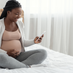 Excited Black Pregnant Woman Sitting With Smartphone On Bed At Home stock photo